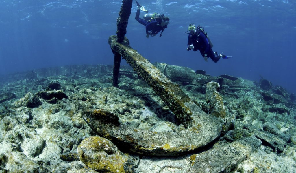 Two divers swimming over a large anchor on the wreck of a ship on the seabed -