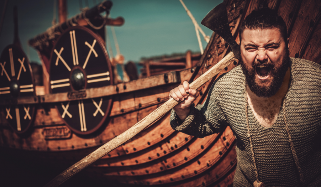 Viking with axe standing near drakkar on the seashore.