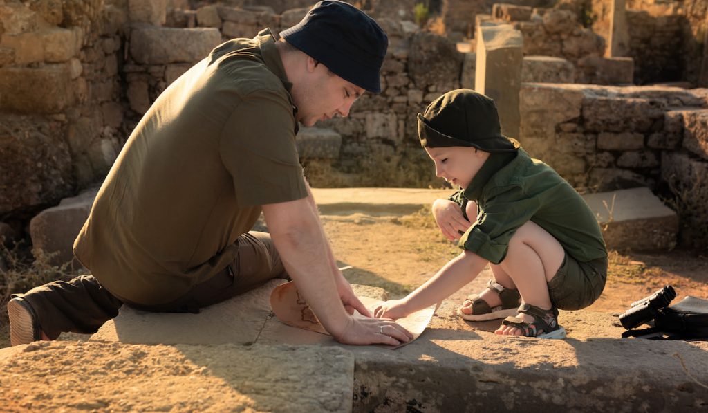 Two archeologists, child and adult, in khaki clothes sitting near ruins and studying an ancient map.
