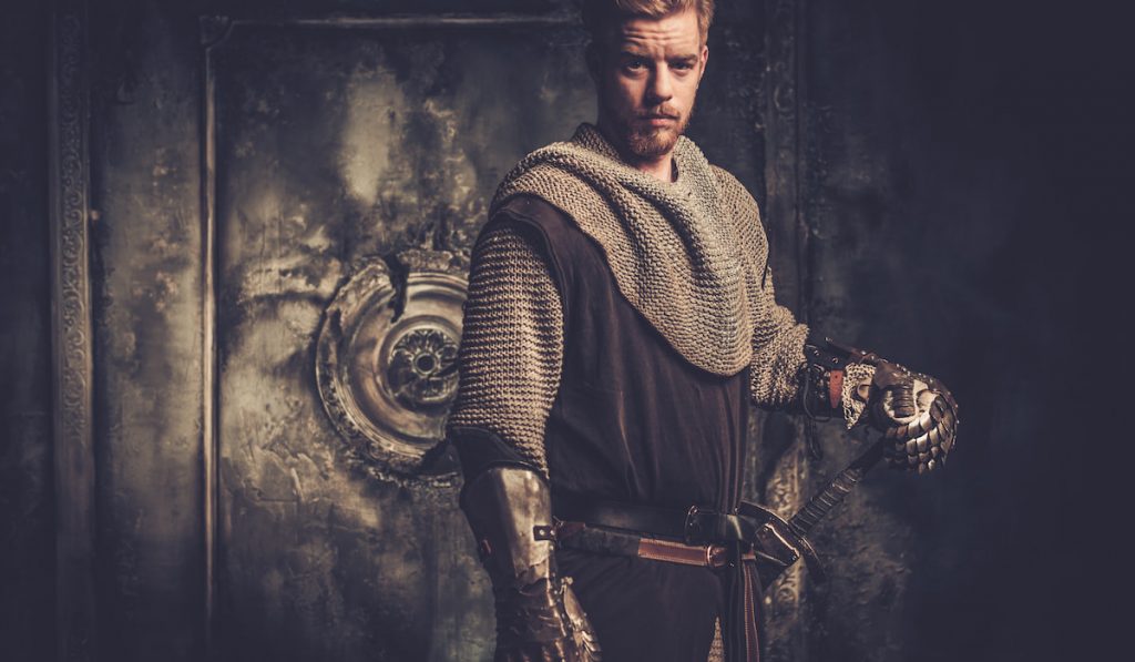 Young medieval knight standing on dark background.
