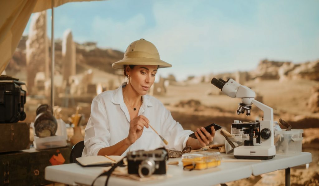 Female Archaeologist Doing Research, Uses Smartphone  and camera to Share Discovery of Fossil Remains, Ancient Civilization Cultural Artifacts